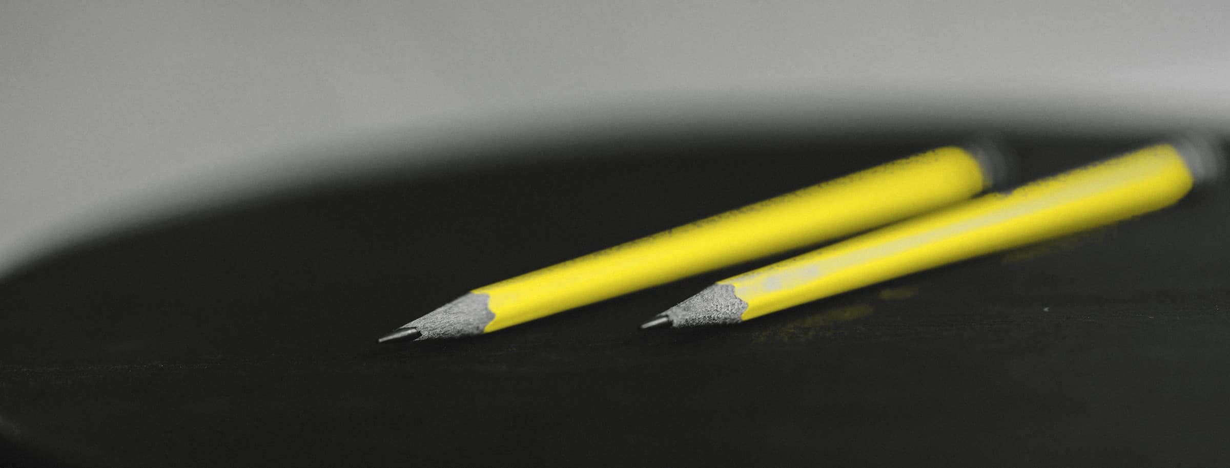 Two sharpened yellow pencils lying on a black surface against a grey wall.